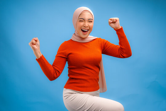 Cheerful young Asian Muslim woman in hijab doing a successful win gesture with clenched fist.