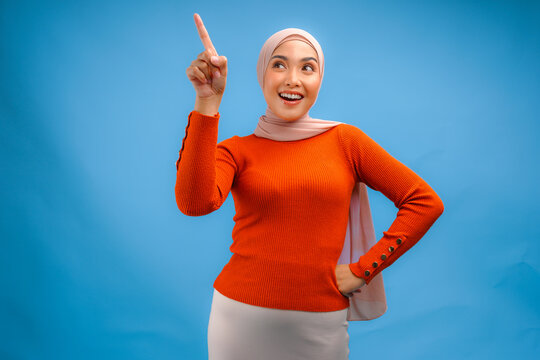 Joyful young Asian Muslim woman in hijab pointing to the copy space upwards.