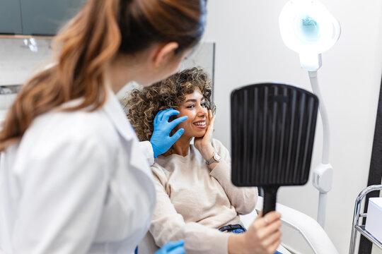 Consultation at aesthetic clinic. Woman looking in mirror. Healthcare and medicine concept. Female doctor in white coat examining patient face after cosmetic procedure or treatment in bright office.