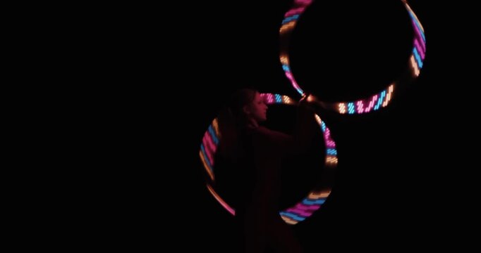 A circus female performer spins LED hoops in the dark, creating bright neon shapes from light trails against the black background of the studio. Slow motion.