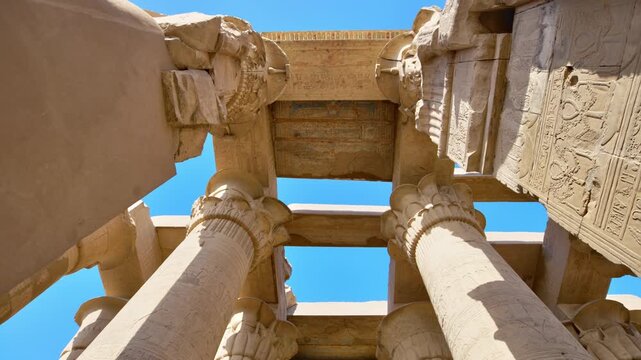 Kom Ombo temple columns with composite capitals and hieroglyphic carvings viewed from below against blue sky in Egypt, upward perspective of ancient sandstone architecture.