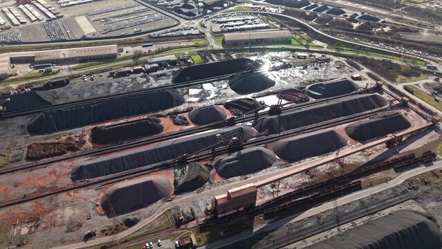Aerial view of dark, textured piles of coal and ore contrasting against the red-stained ground and industrial structures under a bright sky, Immingham, England, United Kingdom.