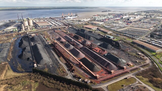 Aerial view of dark, imposing mounds of coal and raw materials sit in stark contrast to the industrial port and distant river, Immingham, England, United Kingdom.