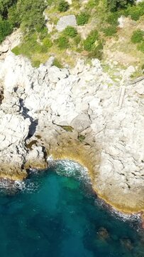 Aerial view of rugged rocky cliffs and clear turquoise water along the Capo di Conca coastline, surrounded by lush greenery. The scene captures the natural beauty and dramatic landscape of the Amalfi