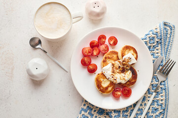 Savory syrniki (cheese pancakes) topped with sour cream, crispy fried onions, and fresh cherry tomatoes, served on a white plate with a blue knit cloth. Perfect for breakfast or brunch.