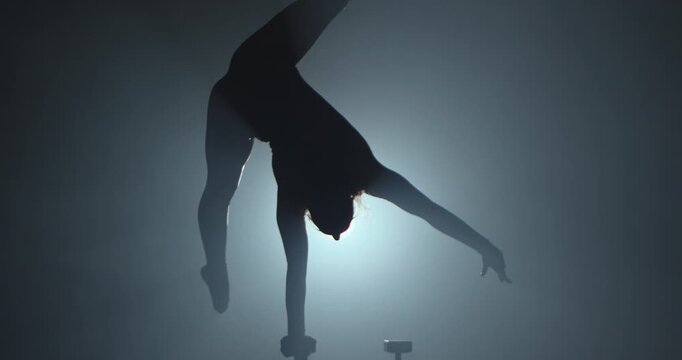 Full shot silhouette of a young circus gymnast performs a arms handstand on canes in a dark studio against a spotlight. For acrobatics, circus show and video production. Slow motion.