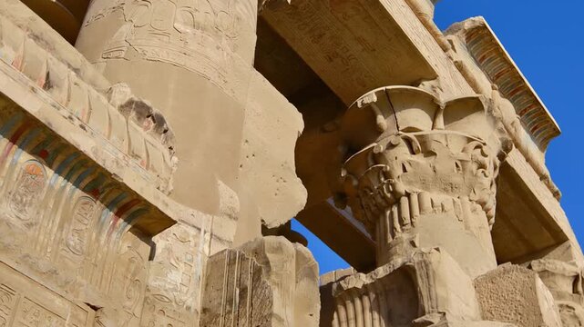 Kom Ombo temple columns with composite capitals and hieroglyphic carvings viewed from below against blue sky in Egypt, upward perspective of ancient sandstone architecture.