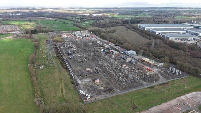 Aerial view of a sprawling electrical substation complex, where metallic structures contrast against the muted green fields, Birmingham, United Kingdom.