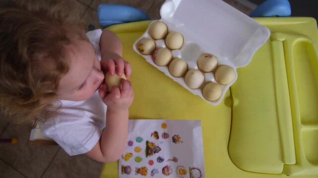 Moscow, Russia, 12.04.2025 Little caucasian girl holding an easter egg and applying a sticker. Kid decorating paschal eggs for easter celebration. Happy spring holiday craft activity.