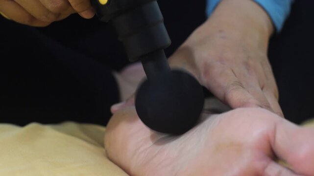 Anonymous woman massaging her foot with a portable electric massage gun at home. Close-up of female foot receiving percussion therapy for pain relief and muscle recovery