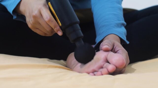 Anonymous woman massaging her foot with a portable electric massage gun at home. Close-up of female foot receiving percussion therapy for pain relief and muscle recovery