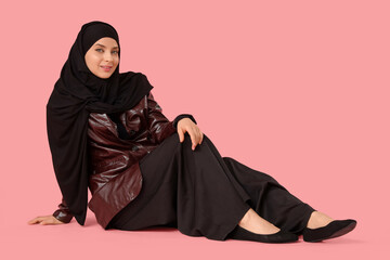 Beautiful young happy stylish Muslim woman in hijab sitting on pink background