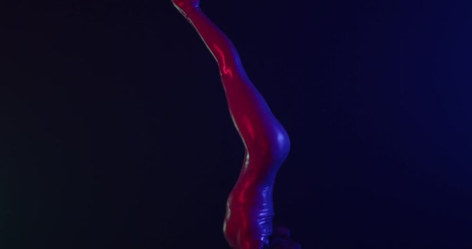 Female acrobat in a shiny bodysuit performs a dramatic handstand split on a pedestal, illuminated by vibrant blue and magenta studio lights on a dark stage. Slow motion.