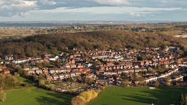 Aerial view of rooftops nestled below a dense, dark forest, softened by the fading sunset glow over Baddesley Ensor, Atherstone, United Kingdom.