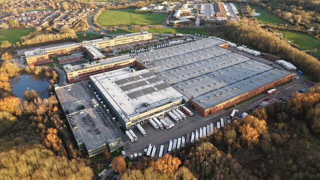 Aerial view of a sprawling industrial complex, where rows of trucks align, contrasting with the surrounding autumnal foliage, Atherstone, United Kingdom.