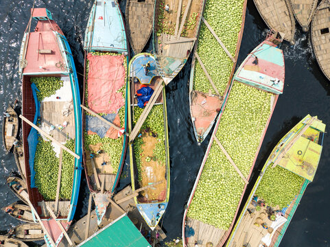 Dhaka, Bangladesh - 23 March 2025: Aerial view of boats laden with vibrant green produce float along the dark, rippling waters near the bustling riverfront.