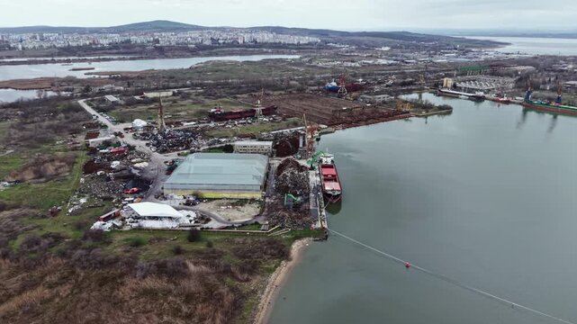 An aerial view of an industrial area beside a river. Ships are docked while construction is ongoing nearby. The surroundings show discarded materials and open land.