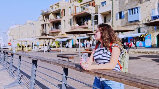 A thoughtful young woman standing by the railing on the wooden boardwalk, with Jaffa's historic