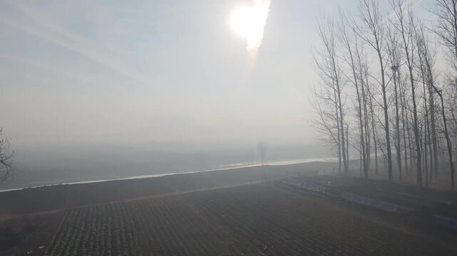 A serene 1080p aerial shot of the bright morning sun shining through a thick mist over vast agricultural fields and a line of bare trees in rural Northern China. March 4, 2026.
