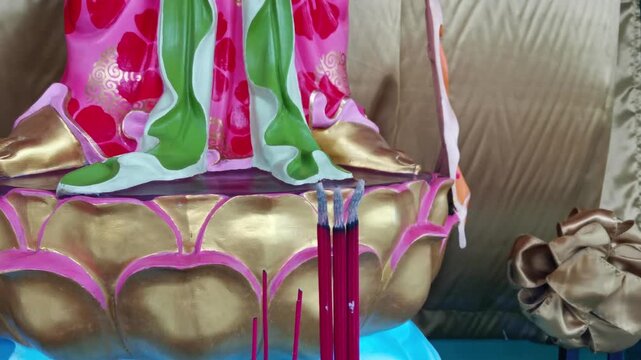 Close-up of red incense sticks standing before a richly painted Chinese deity statue with gold lotus base symbolizing prayer devotion ritual and spiritual offerings