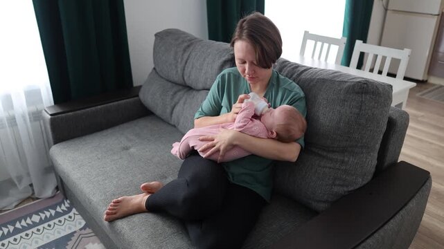High angle shot of a woman sitting on a couch and bottle-feeding a calm baby in a pink romper