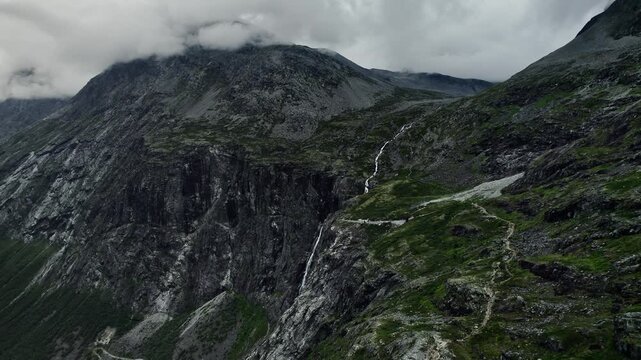 Drone descending near steep cliffs showcasing Trollfossen waterfall and Storgrovfjellet mountain. Clouds cover the peaks as the path leads through the rugged terrain in the natural landscape of Norway