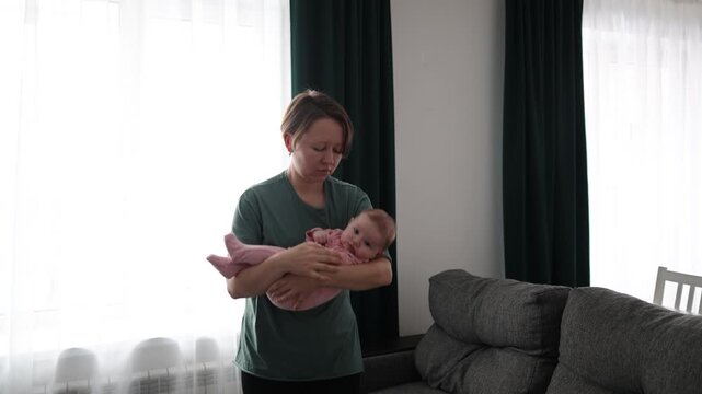 A woman stands in a living room by a window, holding a baby in a pink romper
