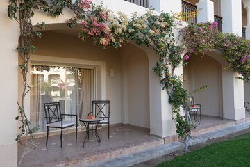 Fototapeta premium Outdoor patio area with two black metal chairs and a small table adorned with flowers and greenery, featuring a glass door leading to an interior space