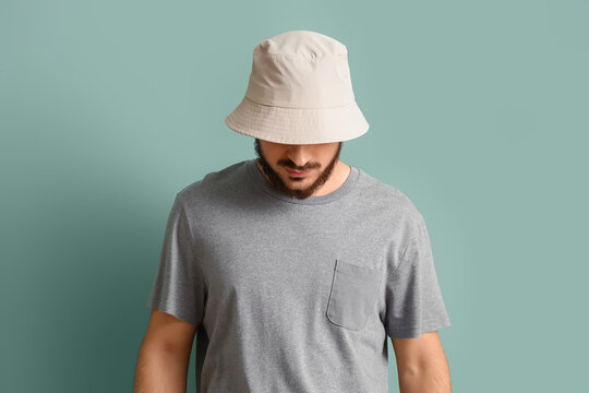 Young bearded man in blank bucket hat and gray t-shirt looking down on pastel blue background, casual streetwear fashion mockup with copy space