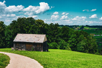 Obraz premium Weathered barn rests on lush green hillside. Curved gravel path invites quiet exploration. Vast blue sky with soft white clouds above. Trees frame the scene, adding depth and calm