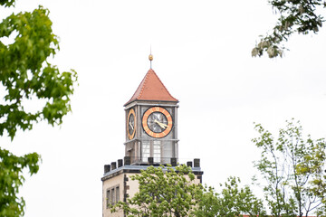Classic clock tower with a red peaked roof and circular dials against a bright sky, framed by green tree branches. Historic urban architecture and heritage.