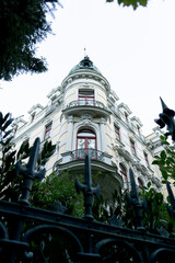 Historic corner building with round tower and ornate balconies viewed from below through decorative fence and foliage. Elegant classical architecture and urban heritage facade. © VladyslavShcherbakov