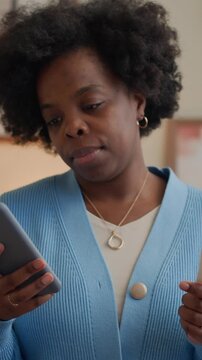 Vertical shot of young African American woman holding pregnancy test and using smartphone in living room
