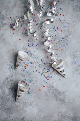 Obraz premium Silver confetti swirls fall against a textured grey surface. Colorful glitter dots scatter across the background. Three paper party cones lie scattered nearby