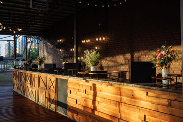Spacious open-air rooftop bar counter crafted from reclaimed wood with geometric patterns, topped with marble, illuminated by hanging Edison string lights and wall sconces against exposed brick walls.