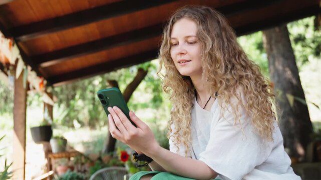 Young woman with long curly hair relaxing on a countryside terrace, scrolling social media on her smartphone during a warm summer day.