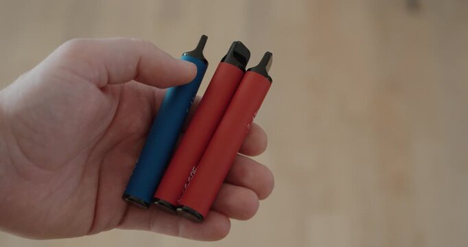 Close-up of a hand holding several disposable e-cigarettes, highlighting health risks, nicotine consumption, and environmental concerns related to electronic waste and recycling.