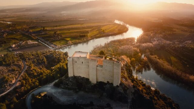 4K cinematic aerial shot of river at sunset over Castle in Miravet, Spain. Historic riverside medieval village with church tower, tiled rooftops and winding river. Golden hour light across countryside