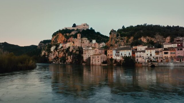 4K cinematic aerial drone of Miravet, Spain at sunset. Cliffside medieval village w historic castle and church above calm river. Warm golden hour light on traditional architecture, dramatic landscape