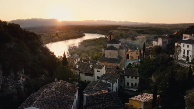 4K cinematic aerial drone shot at sunset over Miravet, Spain. Historic riverside medieval village with church tower, tiled rooftops and winding river. Golden hour light across countryside landscape