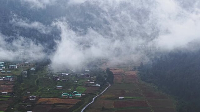 Aerial view of Phobjikha Valley, where mist dances amongst houses and fields, nestled between verdant slopes and a winding path, Nubding, Bhutan.