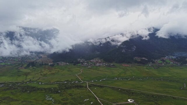 Aerial view of the vast, lush Phobjikha Valley, with mountains shrouded in mist, creating a serene contrast between the green fields and cloudy sky, Nubding, Bhutan.