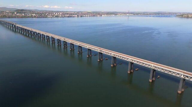 Aerial view of the Tay Rail Bridge spans across tranquil waters, connecting verdant shores under a clear sky, Dundee, United Kingdom.