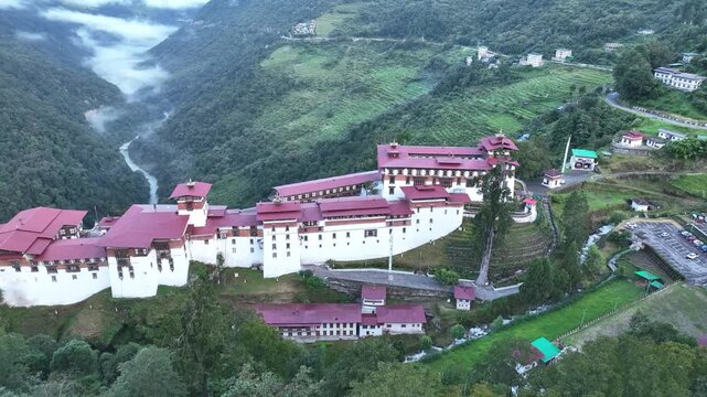 Aerial view of Trongsa Dzong, a fortress with white walls and red roofs, nestled in a lush green valley with a river flowing through it, Trongsa, Trongsa, Bhutan.