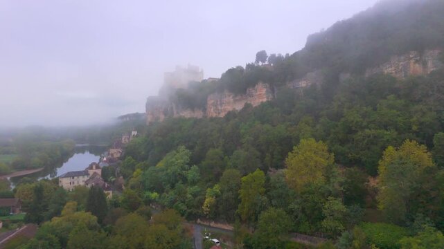 Beynac-et-Cazenac, France - 12 September 2025: Aerial view of Beynac castle perched on a cliff, shrouded in mist, overlooking the river and lush green trees.