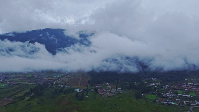 Aerial view of the Phobjikha Valley nestled under a blanket of low-hanging clouds, showcasing the vibrant valley contrasting with the dark, moody sky, Nubding, Bhutan.