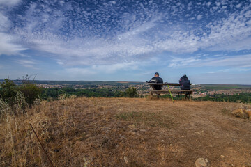 Fototapeta premium Un caminante descansa en un banco de madera junto a su mochila, contemplando un vasto paisaje rural bajo un cielo de nubes dispersas, capturando la esencia del Camino de Santiago