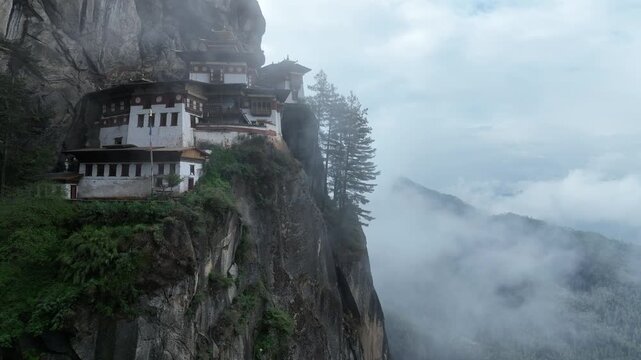 Aerial view of Tiger's Nest Monastery clinging to a cliffside, shrouded in mist, a blend of white walls and dark roofs contrasting with the gray rock, Taktsang, Paro, Bhutan.