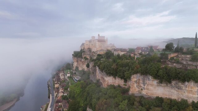 Beynac-et-Cazenac, France - 12 September 2025: Aerial view of Chateau de Beynac perched dramatically atop a cliff, partially veiled in mist, overlooking the tranquil river.