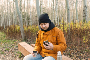Man checking phone while holding cup on bench in birch grove
